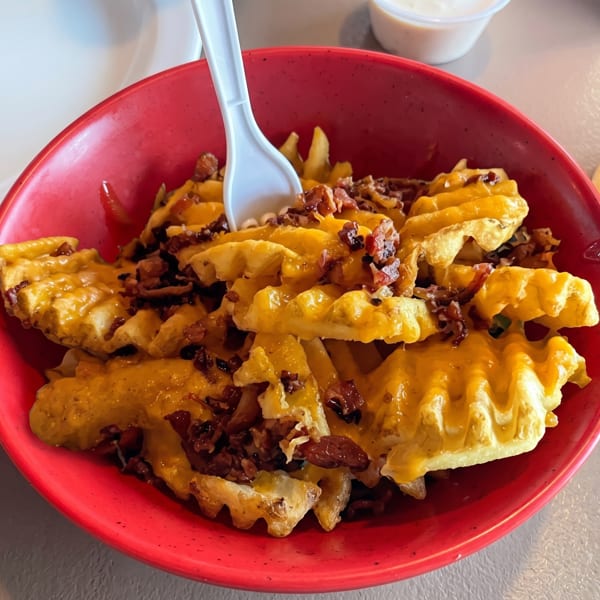 A red bowl filled with waffle fries, melted cheese, and bacon bits, with a white plastic fork.