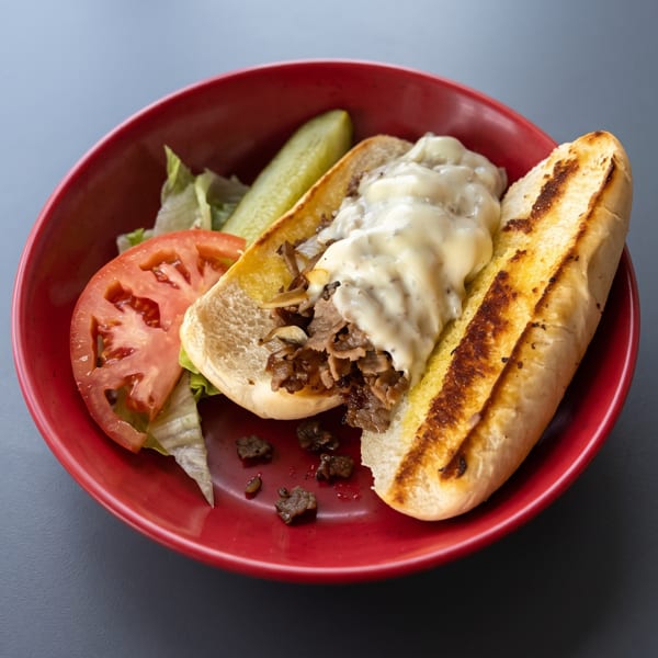 Cheesesteak sandwich with melted cheese, lettuce, tomato, and a pickle on a red plate.